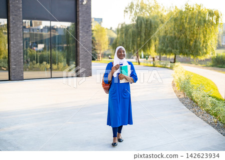 A smiling African American Muslim woman wearing a hijab and a blue dress walks outdoors holding books and a backpack A smiling African American Muslim woman wearing a hijab and a blue dress walks outdoors holding books and a backpack 142623934