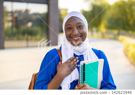 A happy African American Muslim woman wearing a hijab smiles while holding books and a tablet outdoors A happy African American Muslim woman wearing a hijab smiles while holding books and a tablet outdoors 142623946