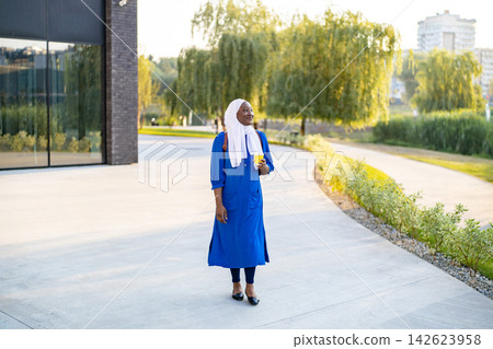 A smiling African American Muslim woman wearing a hijab and a blue dress walks outdoors holding a yellow cup A smiling African American Muslim woman wearing a hijab and a blue dress walks outdoors holding a yellow cup 142623958