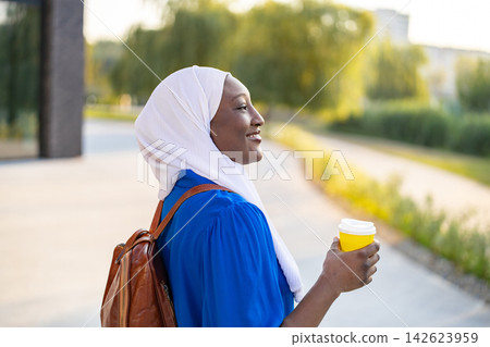 A smiling African American Muslim woman wearing a white hijab and blue top holds a yellow coffee cup while carrying a backpack A smiling African American Muslim woman wearing a white hijab and blue top holds a yellow coffee cup while carrying a backpack 142623959