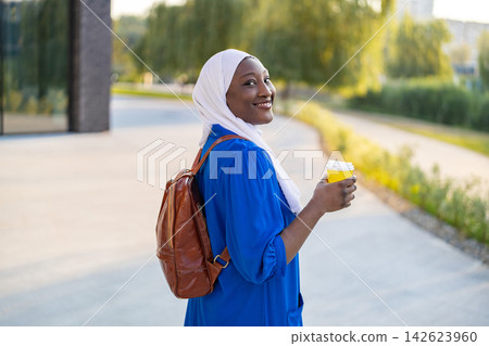 An African American Muslim woman wearing a white hijab and blue dress smiles while holding a coffee cup and carrying a backpack outdoors An African American Muslim woman wearing a white hijab and blue dress smiles while holding a coffee cup and carrying a backpack outdoors 142623960