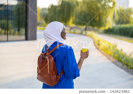 An African American Muslim woman wearing a hijab and a blue dress walks with a backpack and a coffee cup An African American Muslim woman wearing a hijab and a blue dress walks with a backpack and a coffee cup 142623962