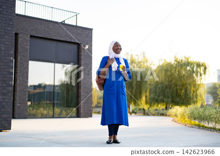 A smiling African American Muslim woman wearing a hijab and a blue dress holds a coffee cup while standing outside a modern building A smiling African American Muslim woman wearing a hijab and a blue dress holds a coffee cup while standing outside a modern building 142623966