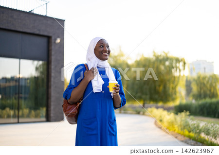 A happy African American Muslim woman wearing a white hijab and blue dress holds a coffee cup while walking outdoors A happy African American Muslim woman wearing a white hijab and blue dress holds a coffee cup while walking outdoors 142623967