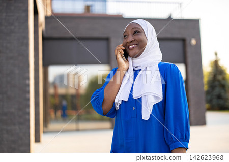 A happy African American Muslim woman wearing a white hijab and blue dress is talking on her smartphone outdoors A happy African American Muslim woman wearing a white hijab and blue dress is talking on her smartphone outdoors 142623968