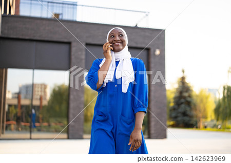 A happy African American Muslim woman wearing a blue dress and white hijab is talking on her mobile phone outdoors A happy African American Muslim woman wearing a blue dress and white hijab is talking on her mobile phone outdoors 142623969