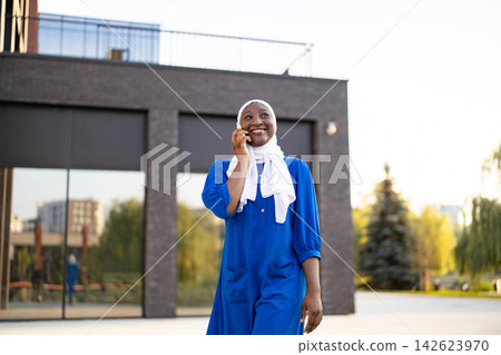 A smiling African American Muslim woman wearing a hijab and blue dress talks on her mobile phone outside a modern building A smiling African American Muslim woman wearing a hijab and blue dress talks on her mobile phone outside a modern building 142623970