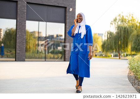 A happy African American Muslim woman wearing a blue dress and white hijab walks outdoors while talking on her smartphone A happy African American Muslim woman wearing a blue dress and white hijab walks outdoors while talking on her smartphone 142623971