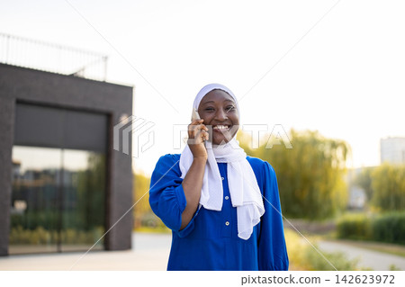A happy African American Muslim woman wearing a hijab and blue dress talks on her mobile phone outdoors A happy African American Muslim woman wearing a hijab and blue dress talks on her mobile phone outdoors 142623972