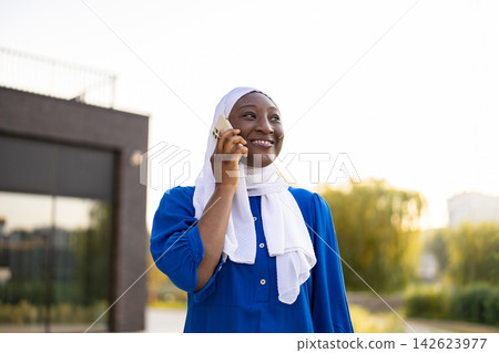 A happy African American Muslim woman wearing a white hijab and blue dress is talking on her mobile phone outdoors A happy African American Muslim woman wearing a white hijab and blue dress is talking on her mobile phone outdoors 142623977