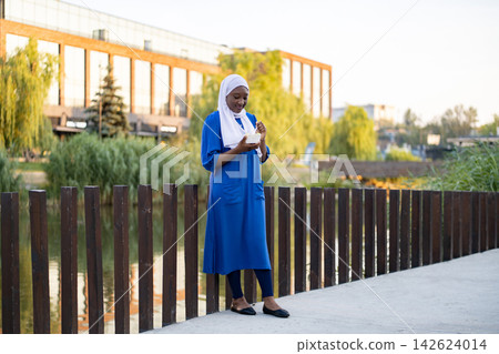 A young African American Muslim woman wearing a hijab and a blue dress enjoys a meal outdoors by a body of water A young African American Muslim woman wearing a hijab and a blue dress enjoys a meal outdoors by a body of water 142624014