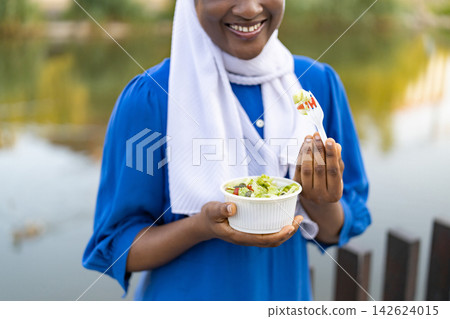 A happy African American woman wearing a white hijab enjoys a healthy salad outdoors by a body of water A happy African American woman wearing a white hijab enjoys a healthy salad outdoors by a body of water 142624015