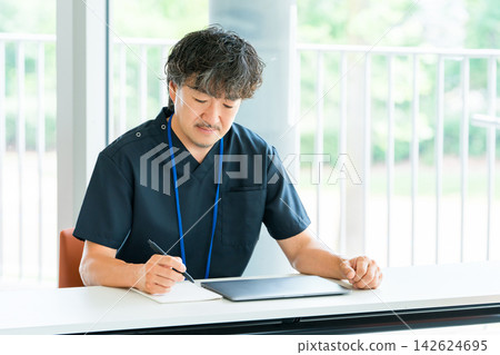 A male medical staff member in scrubs studying while taking notes; a professional engaged in training and skill development. A male medical staff member in scrubs studying while taking notes; a professional engaged in training and skill development. 142624695
