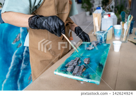 Close-up of female artist arranging textured elements on blue epoxy resin abstract painting. Close-up of female artist arranging textured elements on blue epoxy resin abstract painting. 142624924