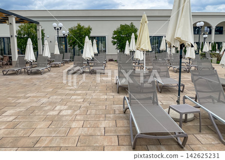 Empty sunbeds and umbrellas on pool terrace at spa resort exterior on cloudy day Empty sunbeds and umbrellas on pool terrace at spa resort exterior on cloudy day 142625231