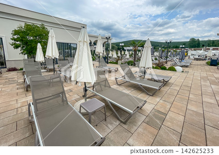 Empty sunbeds and umbrellas on pool terrace at spa resort exterior on cloudy day Empty sunbeds and umbrellas on pool terrace at spa resort exterior on cloudy day 142625233