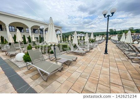 Empty sunbeds and umbrellas on pool terrace at spa resort exterior on cloudy day Empty sunbeds and umbrellas on pool terrace at spa resort exterior on cloudy day 142625234