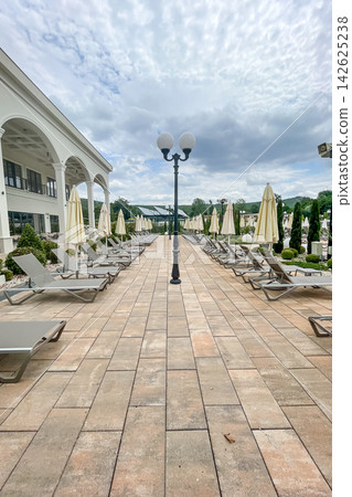 Empty sunbeds and umbrellas on pool terrace at spa resort exterior on cloudy day Empty sunbeds and umbrellas on pool terrace at spa resort exterior on cloudy day 142625238