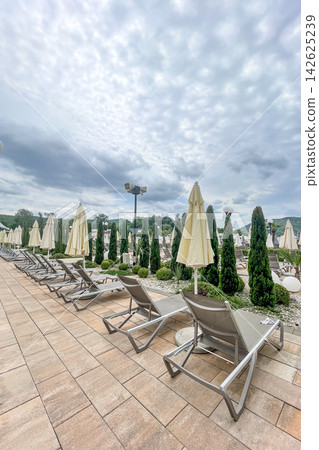 Empty sunbeds and umbrellas on pool terrace at spa resort exterior on cloudy day 142625239
