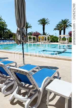 Empty sunbeds and umbrellas on pool terrace at spa resort exterior on cloudy day Empty sunbeds and umbrellas on pool terrace at spa resort exterior on cloudy day 142625240