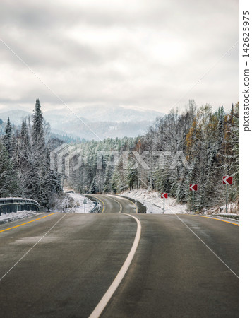 Beautiful empty highway road with road signs in winter pine forest. Snowy hills and mountains under cloudy sky. Winter asphalt road. Vertical scenic nature background with copy space. Seasonal travel Beautiful empty highway road with road signs in winter pine forest. Snowy hills and mountains under cloudy sky. Winter asphalt road. Vertical scenic nature background with copy space. Seasonal travel 142625975