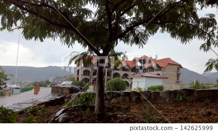 residential complex with red roofs behind tree crown against mountains residential complex with red roofs behind tree crown against mountains 142625999