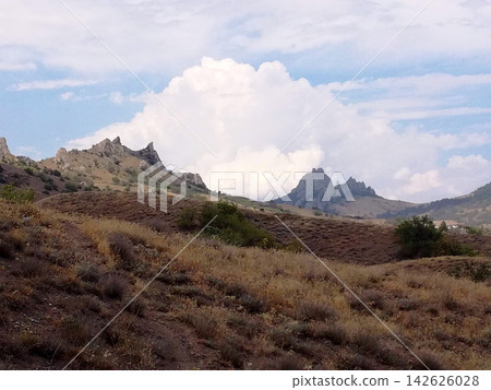 a hilly valley covered in dry grass with jagged mountain peaks under a huge white cloud 142626028