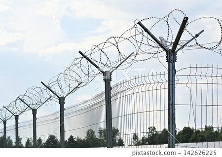 Close up of barbed wire stretched atop steel fence against sky on summer day. Close up of barbed wire stretched atop steel fence against sky on summer day. 142626225