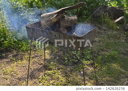 Grill with large firewood pieces and rising smoke in garden. Metal grill on thin legs holding burning logs, outdoor barbecue equipment on grass, rustic cooking scene, summer lifestyle, natural 142626236