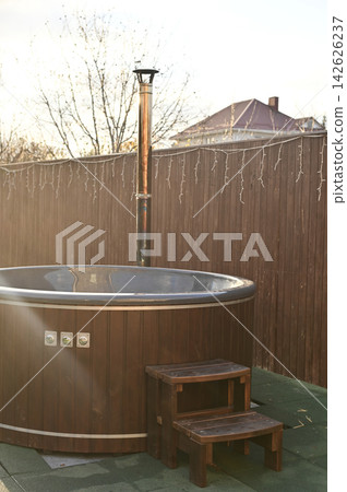 vertical photo, hot tub designed as round wooden barrel on terrace beside wall Outdoor. Hot tub prepared for hydro massage and rest, hot tub symbolizing comfort, leisure and wellness in private yard, vertical photo, hot tub designed as round wooden barrel on terrace beside wall Outdoor. Hot tub prepared for hydro massage and rest, hot tub symbolizing comfort, leisure and wellness in private yard, 142626237