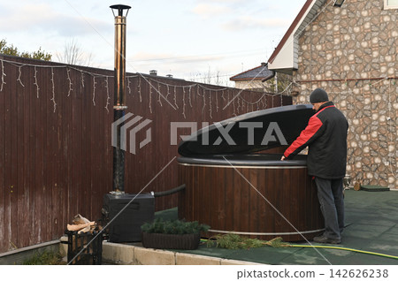 Adult man checks wooden hot tub in backyard garden. Outdoor bath with stove stands near brown fence. Man prepares spa equipment jacuzzi for relaxation. Round wood fired tub looks clean and ready. Adult man checks wooden hot tub in backyard garden. Outdoor bath with stove stands near brown fence. Man prepares spa equipment jacuzzi for relaxation. Round wood fired tub looks clean and ready. 142626238
