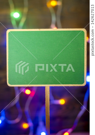 Blank green wooden sign against dark, blurry background with colorful Christmas bokeh lights for custom text or holiday greetings. Template copy space or mockup for celebration or festival background 142627013