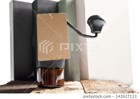 Manual hand coffee grinder with a blank kraft paper tag, resting on rustic bricks with books behind. Template for copy space or mockup Manual hand coffee grinder with a blank kraft paper tag, resting on rustic bricks with books behind. Template for copy space or mockup 142627113