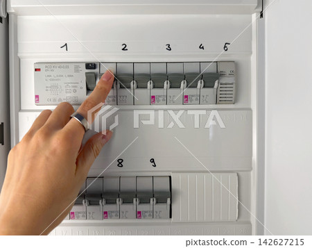 Close-up of a hand checking circuit breakers in a residential electrical distribution panel with multiple labeled switches and protective devices. Close-up of a hand checking circuit breakers in a residential electrical distribution panel with multiple labeled switches and protective devices. 142627215