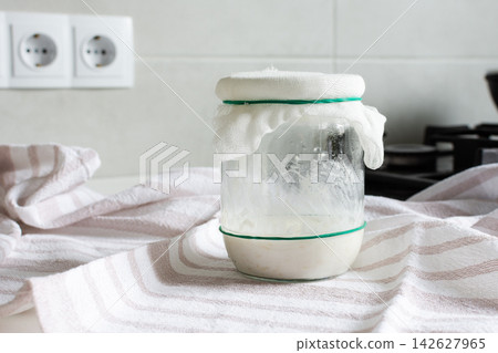 A jar with gluten-free sourdough starter sits on a kitchen counter covered with a cloth. The kitchen has light colors and modern appliances nearby, showing active bread preparation A jar with gluten-free sourdough starter sits on a kitchen counter covered with a cloth. The kitchen has light colors and modern appliances nearby, showing active bread preparation 142627965