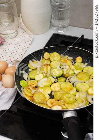 Fried zucchini is cooked with scrambled eggs in a pan on a stovetop. The scene shows a kitchen preparing a meal. Fresh ingredients are visible, creating a simple dish Fried zucchini is cooked with scrambled eggs in a pan on a stovetop. The scene shows a kitchen preparing a meal. Fresh ingredients are visible, creating a simple dish 142627969