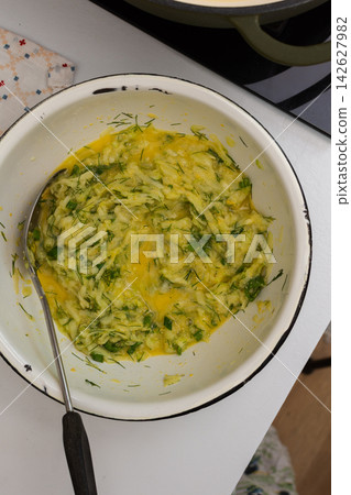 A person is mixing grated zucchini, herbs, and eggs in a bowl in a kitchen. Ingredients like flour are nearby. This is happening during meal preparation time A person is mixing grated zucchini, herbs, and eggs in a bowl in a kitchen. Ingredients like flour are nearby. This is happening during meal preparation time 142627982