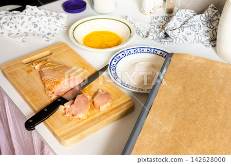 Fresh chicken is cut into pieces while a plate of flour and a bowl of batter are prepared for breading. Cooking happens in a cozy kitchen space Fresh chicken is cut into pieces while a plate of flour and a bowl of batter are prepared for breading. Cooking happens in a cozy kitchen space 142628000
