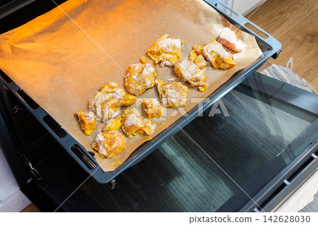 Hands hold a baking tray with deep-fried chicken nuggets covered in flour. The scene is set in a home kitchen with light colors and a casual atmosphere 142628030
