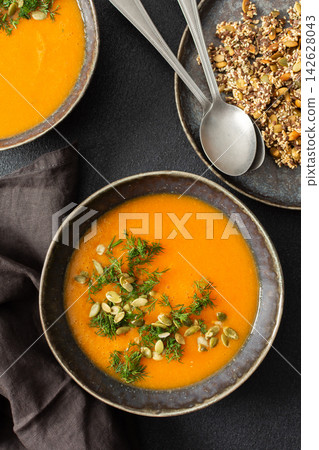 A bowl of pumpkin soup is set on a dark surface. The soup has a smooth texture and is garnished with seeds and herbs. This dish is suitable for breakfast or lunch A bowl of pumpkin soup is set on a dark surface. The soup has a smooth texture and is garnished with seeds and herbs. This dish is suitable for breakfast or lunch 142628043
