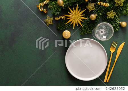 A Christmas table setting with an empty plate and cutlery on a green background. Spruse branches and golden tree decorations add elegance and festive cheer to the scene A Christmas table setting with an empty plate and cutlery on a green background. Spruse branches and golden tree decorations add elegance and festive cheer to the scene 142628052