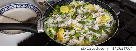 Cooking gluten-free pasta with fried zucchini, peas, and green beans in a skillet on a kitchen stove. Bowl and plate are nearby. It is mid-afternoon, banner Cooking gluten-free pasta with fried zucchini, peas, and green beans in a skillet on a kitchen stove. Bowl and plate are nearby. It is mid-afternoon, banner 142628064