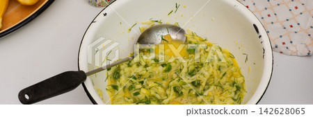 A person is mixing grated zucchini, herbs, and eggs in a bowl in a kitchen. Ingredients like flour are nearby. This is happening during meal preparation time, banner A person is mixing grated zucchini, herbs, and eggs in a bowl in a kitchen. Ingredients like flour are nearby. This is happening during meal preparation time, banner 142628065
