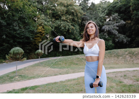 Young woman exercising with dumbbells in a park, promoting healthy lifestyle and fitness 142628339