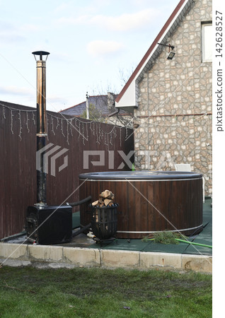 vertical photo, Metal stove positioned near round wooden hot tub on patio. Stove connected by pipe for heating water. stove creating cozy outdoor spa atmosphere, wellness lifestyle concept, 142628527