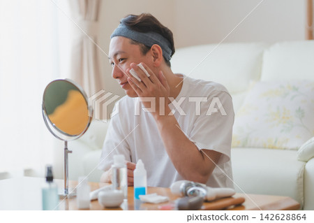 一位四十多歲的男子對著擺滿化妝品的桌上鏡子,用化妝棉輕拍臉頰——這是男士的美容程序。 一位四十多歲的男子對著擺滿化妝品的桌上鏡子,用化妝棉輕拍臉頰——這是男士的美容程序。 142628864