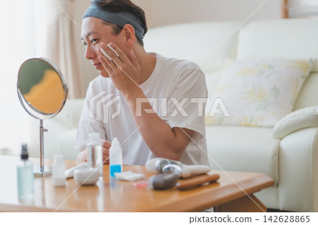 一位四十多歲的男子對著桌上的鏡子,鏡子前擺放著化妝品,他用化妝棉輕拍臉頰,這是他日常男士美容程序的一部分。 一位四十多歲的男子對著桌上的鏡子,鏡子前擺放著化妝品,他用化妝棉輕拍臉頰,這是他日常男士美容程序的一部分。 142628865
