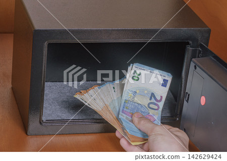 Hand holding euro banknotes depositing cash into a black safe with an open door, interior view showing a felt-lined compartment and wooden surface in the background Hand holding euro banknotes depositing cash into a black safe with an open door, interior view showing a felt-lined compartment and wooden surface in the background 142629424