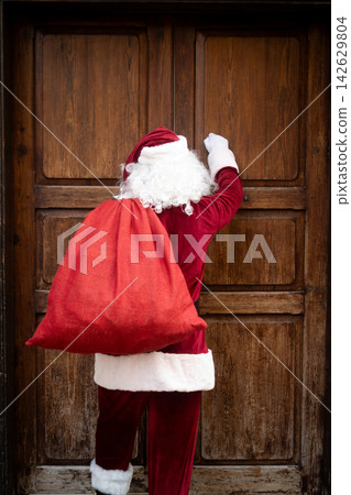 Santa Claus knocking on wooden door with Christmas gift sack Santa Claus knocking on wooden door with Christmas gift sack 142629804