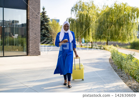A smiling African American Muslim woman in a blue hijab and dress walks with a yellow suitcase while looking at her phone A smiling African American Muslim woman in a blue hijab and dress walks with a yellow suitcase while looking at her phone 142630609